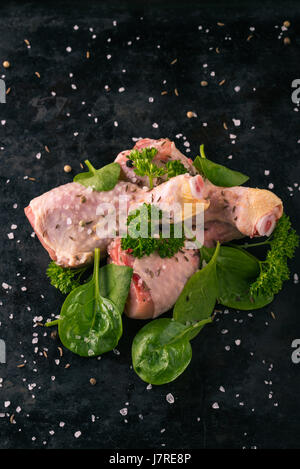 Vertikale Foto von rohem Hühnerfleisch Beine. Drei Stücke von Fleisch sind auf dunkle Vintage Tablett gelegt. Grüne Kräuter wie Petersilie und Spinat sind um mit verschüttet. Stockfoto