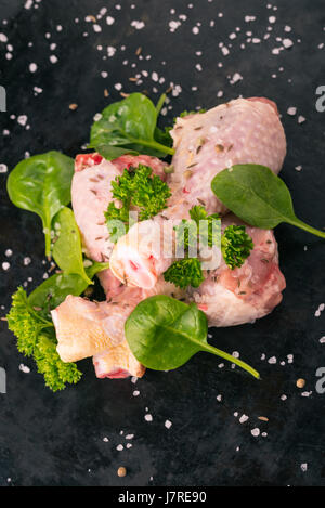 Vertikale Foto von rohem Hühnerfleisch Beine. Drei Stücke von Fleisch sind auf dunkle Vintage Tablett gelegt. Grüne Kräuter wie Petersilie und Spinat sind um mit verschüttet. Stockfoto