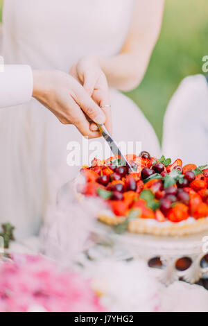 Nahaufnahme Foto von Brautpaar Hände halten das Messer und schneiden das erste Stück der Hochzeitstorte mit Kirschen und Erdbeeren bei der Sonne Stockfoto