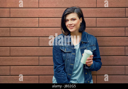 Attraktive Brünette junge Frau mit Tunnel in den Ohren in Blue Jeans Jacke stand vor Ziegel Wand in ihrem heißen Kaffee genießen. Stockfoto