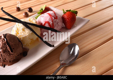 Erdbeere, Vanille und Schokoladeneis Kugeln auf weißen rechteckigen Platte auf Holztisch. Dekoriert mit Obst, Schokokugeln, Vanille auf Ast Stockfoto