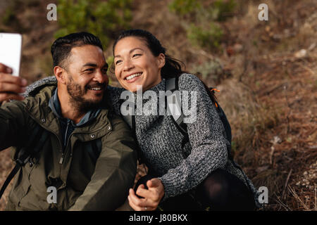 Junges Liebespaar unter Selbstporträt mit ihrem Handy. Mann und Frau sitzen auf Bergweg nehmen Selfie. Stockfoto