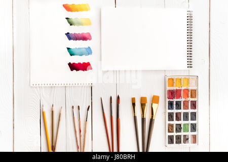 Draufsicht der Reihe von Farben, verschiedene Pinsel, leere Arbeitsmappe und farbenfrohe Pinselstriche auf dem Papier Stockfoto