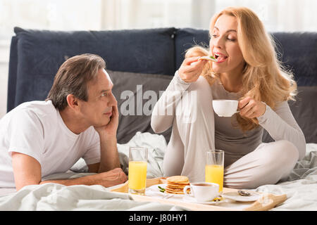 Lächelnde reifer Mann Blick auf schöne Frau essen Pfannkuchen im Bett Stockfoto