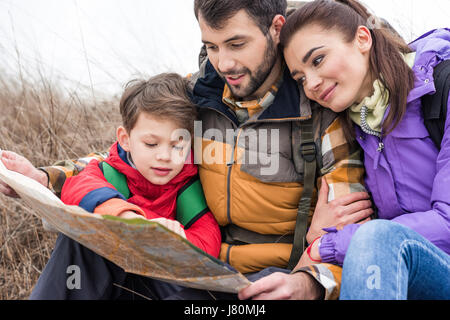 Junge Familie mit Rucksäcken auf dem Rasen sitzen und Blick auf Karte in Landschaft am Herbsttag Stockfoto