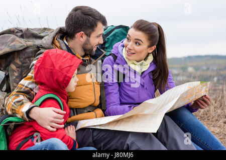 Junge Familie mit Rucksäcken auf dem Rasen sitzen und Blick auf Karte in Landschaft am Herbsttag Stockfoto