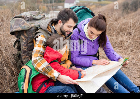 Junge Familie mit Rucksäcken auf dem Rasen sitzen und Blick auf Karte in Landschaft am Herbsttag Stockfoto