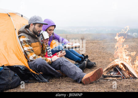 Familie mit einem Kind in der Nähe von Zelt sitzen und trinken heißen Tee aus der Thermoskanne beim Ausruhen in der Nähe von Feuer in Landschaft Stockfoto