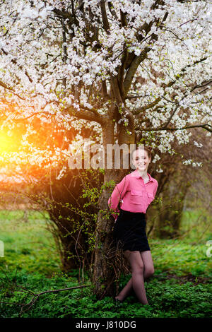 Junge schöne Mädchen in einem rosa Hemd stehen unter blühenden Apfelbaum und genießen einen sonnigen Tag. Blühender Frühling Stockfoto