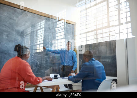 Lächelnden jungen afrikanischen Geschäftsmann hält einen Vortrag an Kollegen stehen vor einer Tafel in einem modernen Büro Stockfoto