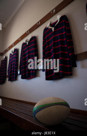 Rugby-Ball auf Bank gegen gestreifte T-shirts hängen in Umkleidekabine Stockfoto