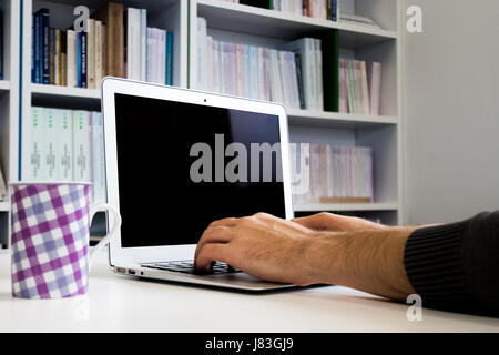 Kerl oder Mann, Student oder Geschäftsmann auf Laptop mit einem leeren Bildschirm und bunte Kaffeetasse auf dem Schreibtisch mit Bücherregal im Hintergrund in l Stockfoto