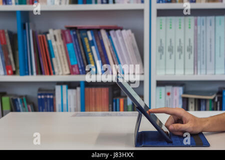 Der Mann arbeitet auf einem Tablet-PC in der Bibliothek mit den Büchern im Hintergrund beim Klicken auf die Anzeige, Foto von der Seite Stockfoto