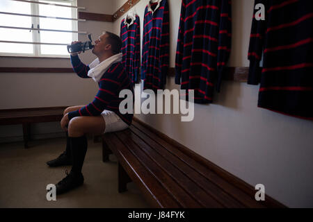 Müde Spieler Trinkwasser sitzend auf Bank gegen Kleiderständer in Umkleidekabine Stockfoto