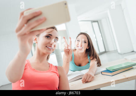 Hübschen weiblichen Studenten in der Schule sitzt am Schreibtisch und Einnahme von Selbstporträts mit einem Smartphone Stockfoto