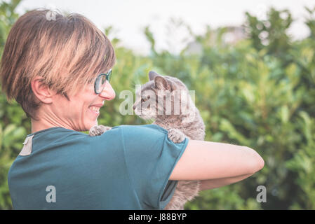 Verspielte Hauskatzen gehalten und gestreichelt von lächelnden Frau mit Brille. Outdoor-Ambiente im grünen Garten. Geringe Schärfentiefe, konzentrierte sich auf anima Stockfoto