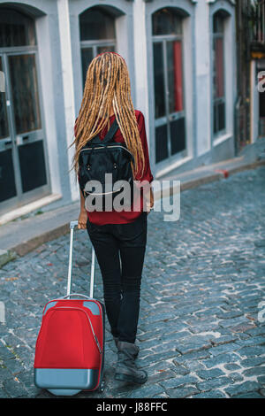 Junge Frau mit blonden Dreadlocks ist auf dem Bürgersteig mit einem roten Koffer. Sicht nach hinten. Reisen und Tourismus. Stockfoto