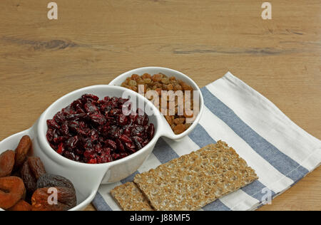 Getrocknete Aprikosen, Cranberries, Rosinen in einer weißen Schüssel und zwei Stücke von Knäckebrot und eine Portion auf einem alten Küchentisch. Enge, horizontale Ansicht. Stockfoto