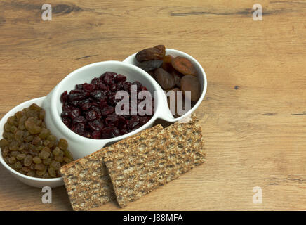 Getrocknete Aprikosen, Cranberries, Rosinen und zwei Stücke von Knäckebrot auf einem alten, hölzernen, Küchentisch. Enge, horizontale Ansicht. Stockfoto