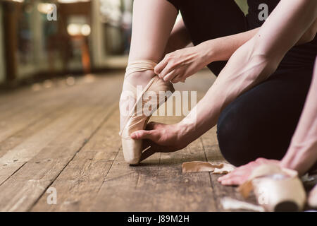 Ballerina oder Tänzerin Mädchen anziehen ihrer Ballettschuhe auf dem Holzboden. Männlichen Balletttänzer hilft Puttiing auf. Close-up. Stockfoto
