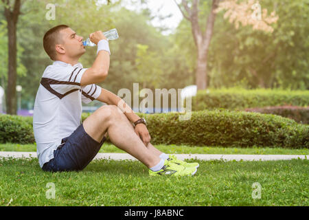 Junge Jogger sitzen auf dem Rasen im Park, von der Morgengymnastik ausruhen und trinken Mineralwasser. Stockfoto