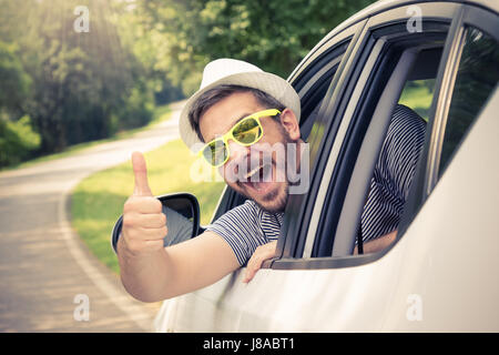 Junger Mann mit Hut und Sonnenbrille Daumen vom Fahrersitz durch geöffnete Fenster auftauchen. Urlaub und Reisen-Konzepte. Stockfoto