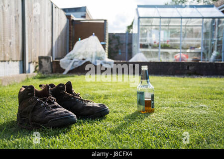 Ein paar Wasser eingeweicht braune Stiefel neben einer offenen Flasche Bier an einem heißen sonnigen Tag. Nach einem harten Arbeitstag im Garten. Gewächshaus im Hintergrund