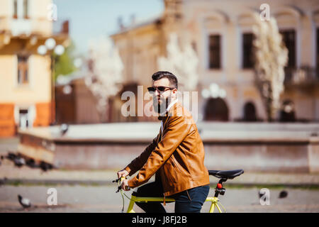 Seitenansicht des gut aussehenden jungen, bärtigen Mann wegschauen während der Fahrt auf seinem Fahrrad im Freien. stilvolle Geschäftsmann mit dem Fahrrad zur Arbeit gehen Stockfoto
