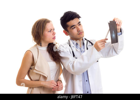 Junge Smartlooking Arzt mit schwarzen Haaren in weißes Kleid mit Stethoskop, Röntgen, junge schöne Frau mit langen braunen Haaren in Beige Kleid auf Stockfoto