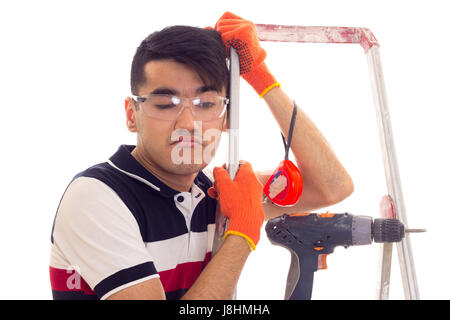 Unglücklich junger Mann mit schwarzen Haaren in weißen und blauen T-shirt und Jeans mit orangefarbenen Handschuhe und Schutzbrille Stand in der Nähe der Ledder mit elektrischem Stockfoto