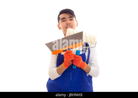 Junger Mann mit dunklen Haaren weißes Hemd und eine blaue insgesamt mit orange Handschuhe mit Spachtel und Roll auf lächelnd weißer Hintergrund im studio Stockfoto