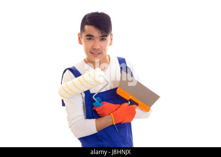 Lächelnder junger Mann mit dunklen Haaren in weißem Hemd und blauen overall mit orange Handschuhe halten Spachtel und Rollen auf weißem Hintergrund im studio Stockfoto