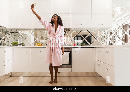 Lächelte, afroamerikanische Frau Selfie in der Küche machen. Frau trägt rosa Bademantel. Stockfoto