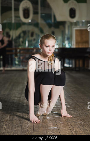 Schöne junge blonde Kaukasische Frau sitzen auf ihren Fuß-Übungen an der Ballettschule. Verschwommene Gruppe von Ballett-Tänzer stehen im Hintergrund Stockfoto