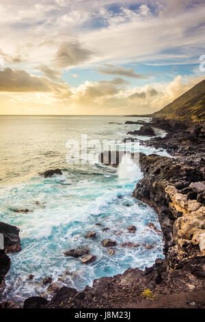 Wellen in die felsige Küste von West-Oahu auf dem Weg zum Kaena Point, gewärmt durch das Sonnenlicht von einem goldenen hawaiianischen Sonnenuntergang. Stockfoto