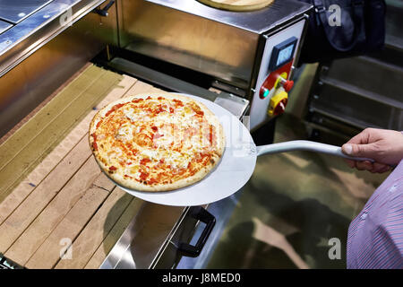 Zubereitete Pizza auf einem Metallspatel wird vom Förderband genommen. Stockfoto