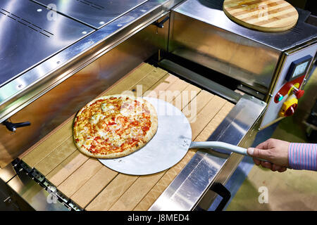 Zubereitete Pizza auf einem Metallspatel wird vom Förderband genommen. Stockfoto