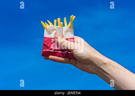 Frisch duftende appetitlich knusprigen Pommes frites in einer Papiertüte in eine männliche Hand auf einem klaren blauen Himmelshintergrund. Stockfoto