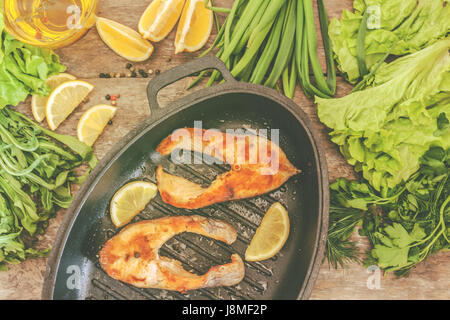 Gebackene Steak Forellen Fischen in gusseisernen Form, rund um die Grüns, Blätter, Salat, Zitrone, Olivenöl, Pfeffer, Gewürze. Von oben, Nahaufnahme. Konzept der healt Stockfoto