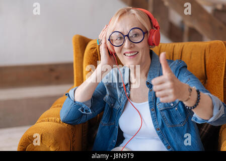 Schöne Auswahl. Attraktive Reife Blondine Lächeln auf ihrem Gesicht zu halten und die Kopfhörer auf dem Sessel sitzend rechts aufsetzen Stockfoto