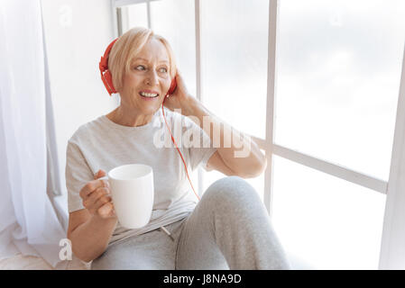 Aktive Musik anhören. Ziemlich lächelnden Frau mit Schale in der rechten Hand, auf der Fensterbank sitzend und auf der Suche zur Seite Stockfoto