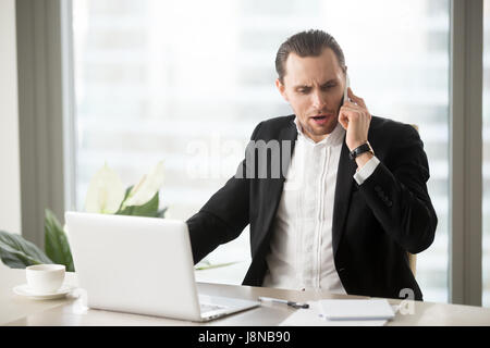 Wütend Geschäftsmann auf dem Handy am Schreibtisch im Büro sprechen. Betonte Unternehmer streiten am Telefon, mit schlechten Nachrichten, unangenehm überrascht h Stockfoto