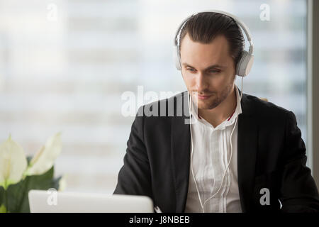 Junger Mann im Kopfhörer hören Musik während der Arbeit am Laptop im Büro, anschauen von Videos, live-Stream, Webinar, erfreut sich Web-Inhalte. Unternehmer nehmen Stockfoto