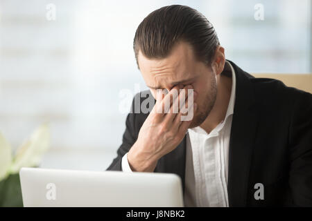 Frustriert Geschäftsmann fühlt Schmerz in den Augen, weil Sehkraft nach langer Arbeit am Computer überfordern. Müde junger Mann massiert Augen vor Laptop. Stockfoto