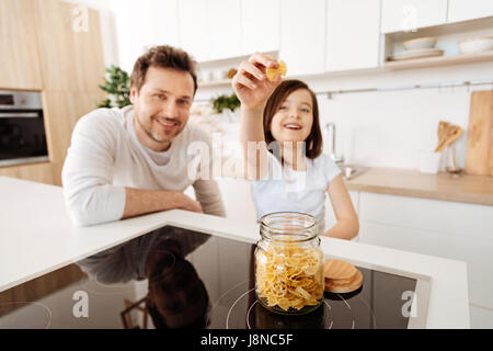 Pasta-Liebhaber. Ziemlich niedlich Mädchen in der Küche Stand und halten ein Stück von Farfalle Nudeln aus dem Glas auf das Kochfeld platziert, während ihr Vater lookin Stockfoto