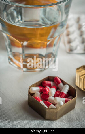 Metallische Vintage Pillbox mit weißen und roten Kapseln zusammen mit Wasserglas Stockfoto