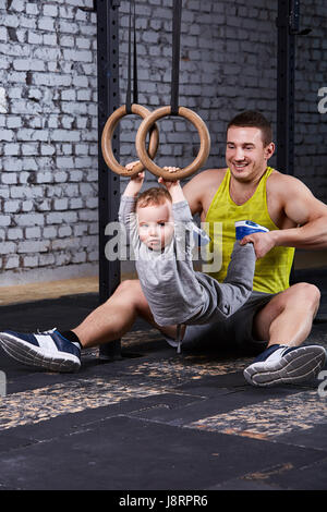 Lächelnd junger Vater Züge der kleine Sohn mit Gimnastic Ringen gegen Ziegel Wand in der Turnhalle. Stockfoto