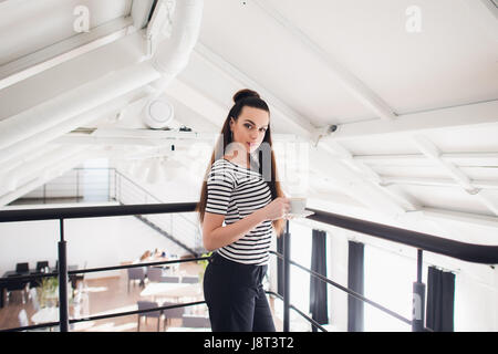 Jung und erfolgreich. Attraktive junge Frau in intelligente Freizeitkleidung Kaffeetasse halten und Blick in die Kamera beim stehen in der Nähe von Fenster im Büro. Stockfoto