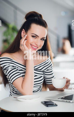 Junge Frau mit einer Tasse Kaffee am Tisch in der Nähe von Tisch sitzen und in die Kamera schaut. Stockfoto