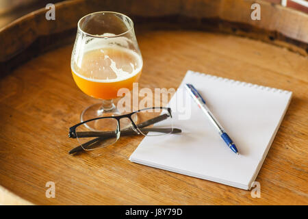 Glas Weizenbier mit ein paar Gläser, Kugelschreiber und Notizblock stehend auf einem Holzfass in einer Brauerei Stockfoto